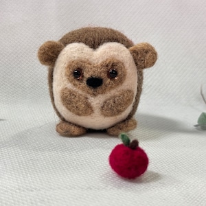 May include: A small, brown and white needle felted hedgehog with black eyes and a brown nose. The hedgehog is sitting on a white surface with a red felt apple in front of it.