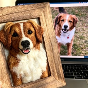 May include: A three-dimensional needle felted portrait of a brown and white dog with floppy ears, black eyes, and a pink tongue, mounted inside a rustic wooden frame. The dog has a white blaze on its face and a fluffy white chest.
