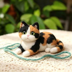 May include: A needle-felted calico cat figurine with black, orange, and white fur patches, green eyes, and a small pink nose. The cat is in a resting position with a piece of teal yarn wrapped around its body on a textured white surface.