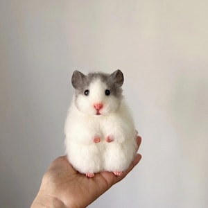 Könnte beinhalten: Eine kleine, flauschige, nadelgefilzte Hamsterfigur mit weißem Körper sowie grauen Ohren und Kopfzeichnung. Der Hamster hat schwarze Perlenaugen, eine kleine rosa Nase und winzige rosa Pfoten und steht aufrecht in einer runden Haltung.