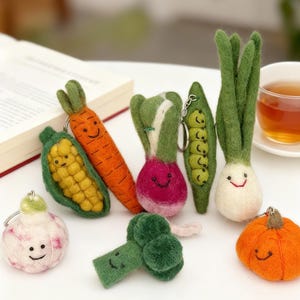 Puede incluir: Una colección de llaveros de fieltro hechos a mano con forma de verduras, incluyendo una zanahoria, maíz, rábano, vaina de guisantes, cebolla, brócoli y calabaza. Cada llavero presenta una cara sonriente y colores vibrantes.