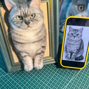 May include: A three-dimensional needle felted gray tabby cat sculpture mounted inside a blue and gold ornate picture frame, positioned next to a smartphone displaying a photograph of the same cat on a white background.
