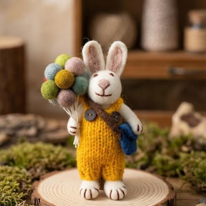 Peut inclure: Figurine de lapin en feutre fait main, vêtu d'une salopette jaune tricotée et tenant un bouquet de ballons en feutre colorés. Le lapin porte une sacoche marron et se tient sur une tranche de bois, un objet décoratif charmant.