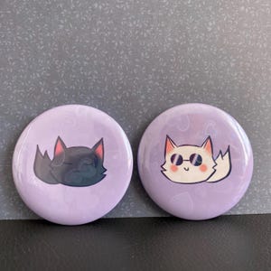 May include: Two round purple buttons with white hearts. One button features a black cat with closed eyes. The other button features a white cat with sunglasses and a pink nose.