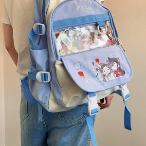 Puede incluir: Una mochila azul claro y crema con múltiples compartimentos y correas ajustables. La solapa frontal presenta una ventana transparente y una colorida ilustración de personajes de anime. La mochila tiene detalles en blanco y azul.