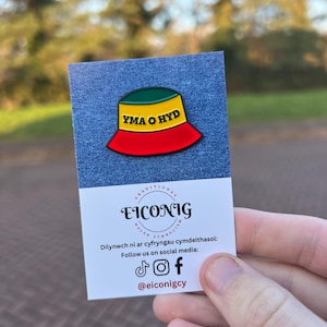 May include: A yellow, green and red enamel pin of a bucket hat with the text "YMA O HYD" on the front. The pin is on a white card with the text "TRADITIONAL WELSH SYMBOLISM EICONIG" and social media handles.