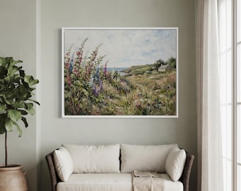 Könnte beinhalten: Ein rechteckiges gerahmtes Landschaftsgemälde mit hohen rosa und blauen Stockrosen im Vordergrund, einem grasbewachsenen Küstenhang und einem kleinen weißen Haus in der Ferne unter einem blassen, bewölkten Himmel, in einem weißen Rahmen.
