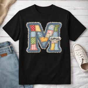Peut inclure: T-shirt noir avec un grand "M" de style patchwork bleu, rempli de carrés de tissu colorés. Le mot "ama" est imprimé dans un petit ovale blanc. Le t-shirt est présenté sur une surface en bois clair avec un jean et une basket blanche.