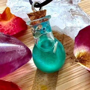 May include: A teal-colored potion bottle pendant with a cork stopper, suspended from a black cord. The bottle is filled with a shimmering liquid. The background includes crystals and flower petals, creating a mystical aesthetic.