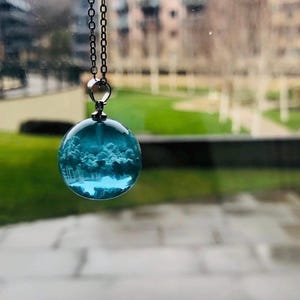 May include: A spherical pendant necklace featuring a clear blue resin orb containing a miniature white cloud formation suspended inside. The pendant is attached to a thin silver-toned metal chain with small circular links, hanging vertically.