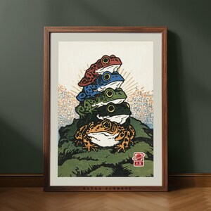 May include: A framed art print featuring a vertical stack of five colourful frogs in red, blue, green, black, and orange, perched on a textured green mound. The background includes a sunburst pattern, and a small red square stamp with text is visible.