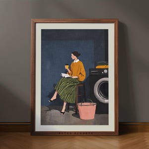 Puede incluir: Una impresión artística enmarcada representa a una persona sentada, leyendo y sosteniendo una taza de café. La persona lleva un suéter amarillo y una falda a rayas verdes. Una lavadora y un cubo rosa están en el fondo. La impresión está enmarcada en un marco de madera oscura.