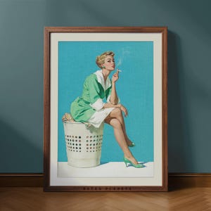 Puede incluir: Una impresión vintage enmarcada que presenta a una mujer con un vestido verde y blanco, sentada en una cesta de lavandería blanca, fumando un cigarrillo. El fondo es de color turquesa. La impresión está en un marco de madera marrón. El texto "RETRO PLANETS" está en la parte inferior.