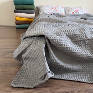 Puede incluir: Una manta de tejido de waffle gris está drapeada sobre una cama con dos almohadas con estampado floral. La manta está doblada al pie de la cama y los bordes son visibles.