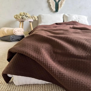 Puede incluir: Una manta marrón texturizada sobre una cama con almohadas blancas. La manta tiene un patrón tejido. Mantas plegadas en beige y gris están apiladas en un taburete pequeño. Un tapiz de macramé está encima de la cama.