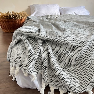 Könnte beinhalten: Eine schwarze und weiße Chevron-Muster-Decke mit weißen Quasten. Die Decke liegt über einem Bett mit weißen Kissen.