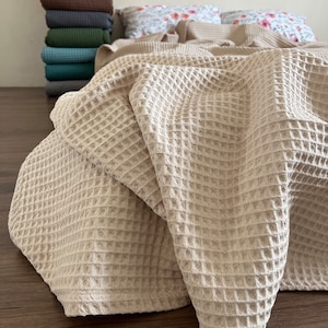 Op de afbeelding: Een beige wafel geweven deken gedrapeerd over een bed met bloemenmotief kussens. De deken is gemaakt van een zachte, textuurrijke stof.
