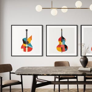 Op de afbeelding: Drie ingelijste prints van abstracte geometrische ontwerpen van muziekinstrumenten. De prints tonen een gitaar, een bas en een elektrische gitaar. De prints hangen aan een witte muur boven een houten tafel met stoelen.