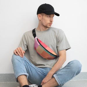 May include: A person wearing a grey t-shirt and blue jeans is sitting down with a black baseball cap on their head. They are wearing a colorful patterned fanny pack across their body.