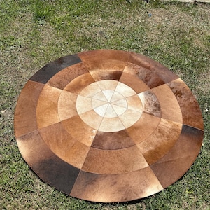 Könnte beinhalten: Ein runder Teppich aus zusammengenähten Rindsleder-Paneelen in verschiedenen Brauntönen, von hellem Beige bis zu dunkler Schokolade. Das Design zeigt ein zentrales Sternmuster, das von konzentrischen Ringen aus tortenförmigen Segmenten umgeben ist.