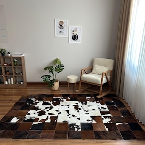 May include: A rectangular cowhide rug with a patchwork design in shades of brown, black, and white. The rug is placed on a wooden floor, with a rocking chair, a small stool, and a potted plant in the background. The room has a neutral color palette.