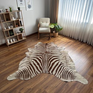 May include: A large, irregularly shaped animal hide rug featuring a brown and cream zebra stripe pattern. The rug is laid flat on a wooden floor, showcasing a natural, organic silhouette with distinct dark brown stripes against a light background.