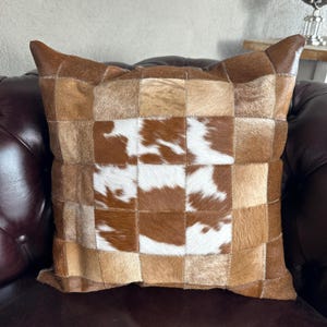 May include: A square cushion with a patchwork design. The cushion has a checkerboard pattern of brown, tan, and white cowhide squares. It is placed on a brown leather sofa.