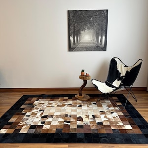 May include: A rectangular cowhide rug with a patchwork design in shades of brown, tan, and white, framed by a black border. The rug is placed on a wooden floor, with a black and white butterfly chair and a small wooden side table nearby.