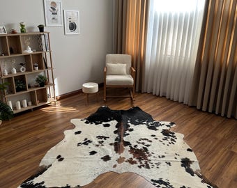 May include: A large, irregularly shaped cowhide rug featuring a natural pattern of white, black, and brown patches. The rug is laid flat on a wooden floor, showcasing its organic, asymmetrical edges and distinct color variations across the surface.