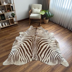 May include: A large, irregularly shaped area rug featuring a brown and cream zebra stripe pattern. The rug is laid flat on a wooden floor, showcasing the natural hide silhouette with distinct dark brown stripes against a lighter cream background.