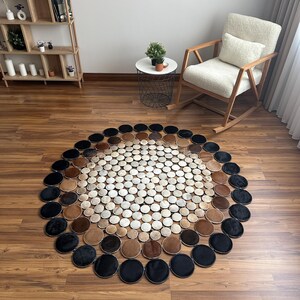 May include: A circular area rug composed of numerous small, stitched circular patches in varying shades of white, tan, brown, and black. The rug features a gradient pattern with lighter colors concentrated in the center and darker colors at the edge.
