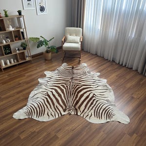 May include: A large, irregularly shaped area rug featuring a brown and white zebra stripe pattern. The rug is laid flat on a wooden floor, showcasing a natural hide-like silhouette with distinct dark brown stripes against a cream-coloured background.