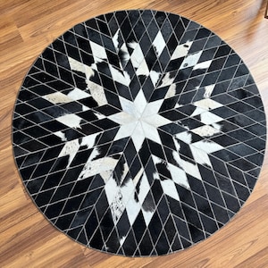 Puede incluir: Una alfombra redonda con un diseño de estrella. La alfombra está hecha de piezas de cuero de vaca en blanco y negro cosidas. El patrón de estrella está centrado, con una estrella blanca en el medio y formas de diamante en blanco y negro irradiando hacia afuera.