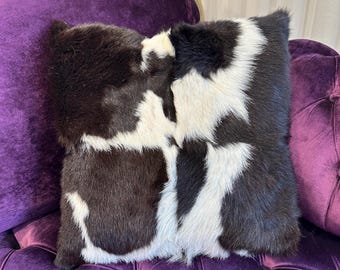 May include: A black and white cowhide pillow with a soft, fuzzy texture. The pillow is sitting on a purple velvet chair.