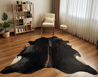 May include: A large, irregularly shaped cowhide rug featuring a deep black base color with subtle brown undertones and distinct white patches along the edges, laid flat on a wooden floor in a room with a wooden bookshelf and a cream-colored chair.