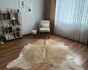 May include: A large, irregularly shaped cowhide rug featuring a natural tan and beige mottled pattern with subtle white highlights, laid flat on a dark brown wood-grain floor in a room with a wooden bookshelf and a cream-colored rocking chair.
