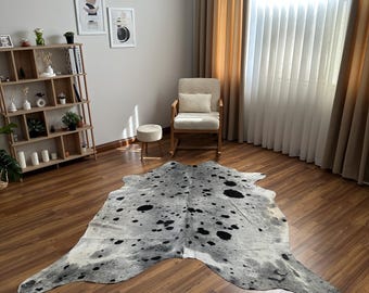 May include: A large, irregularly shaped cowhide rug featuring a mottled pattern of black, gray, and white patches. The rug lies flat on a wooden floor, displaying a natural animal hide texture with distinct dark spots scattered across the surface.