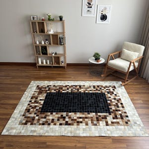 Puede incluir: Una alfombra rectangular con un diseño de patchwork de pequeños azulejos cuadrados en tonos negro, marrón, bronceado y crema. La alfombra tiene un centro rectangular negro sólido rodeado por bordes concéntricos de parches en tonos tierra.