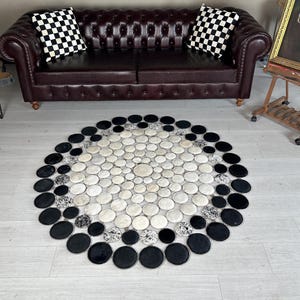 Könnte beinhalten: Ein runder Teppich mit einem Muster aus schwarzen, weißen und gesprenkelten Kreisen. Der Teppich besteht aus einem weichen Material und liegt auf einem hellen Holzboden. Der Teppich liegt vor einem braunen Ledersofa.