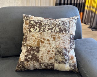 May include: Square decorative pillow with a patchwork design. The pillow features a mix of brown, tan, and white cowhide squares, creating a textured and rustic appearance. The pillow is placed on a gray sofa.