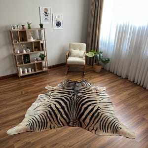 May include: A large, irregularly shaped animal hide rug featuring a distinct zebra-inspired pattern with bold black stripes set against a natural tan and cream-coloured background, laid flat on a polished wooden floor in a room setting.