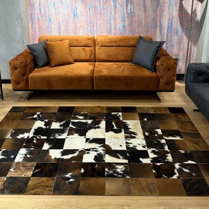 May include: A brown, black, and white cowhide patchwork rug with a checkered pattern. The rug is laid out on a wooden floor in front of a brown sofa with pillows.