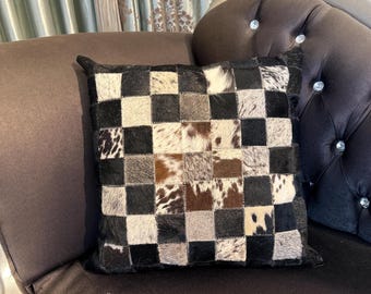 May include: A black and white checkered throw pillow with a patchwork design made from cowhide. The pillow is sitting on a dark brown couch.