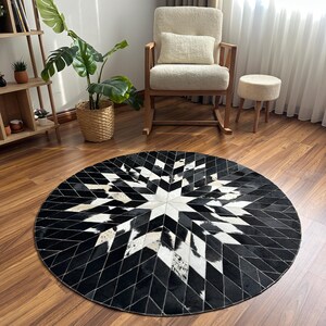 Pode incluir: Um tapete redondo com um design geométrico de estrela. O tapete apresenta retalhos de pele de vaca em preto, branco e branco sujo costurados. O tapete é colocado sobre um chão de madeira, com uma cadeira de balanço e um pequeno banco no fundo.