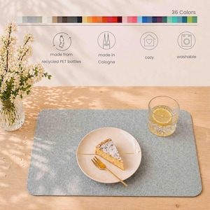May include: A light gray felt placemat with rounded corners, a plate with cake, and a glass of water with lemon. The image also shows a vase of flowers and text that reads "36 Colors", "made from recycled PET bottles", "made in Cologne", "cozy", and "washable".