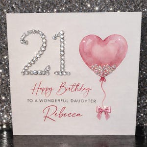 May include: A birthday card with the number "21" embellished with rhinestones. A pink heart-shaped balloon with glitter accents is attached to a pink ribbon and bow. The card reads "Happy Birthday to a wonderful daughter Rebecca."
