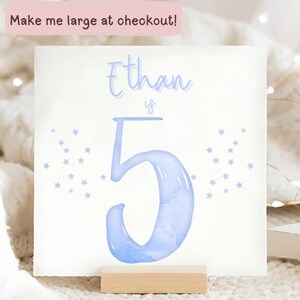 May include: A white square card with the name "Ethan" and the number "5" in light blue watercolor style. The word "is" is between the name and the number. Small blue stars are scattered around the edges. The text "Make me large at checkout!" is at the top.