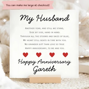 Könnte beinhalten: Eine weiße quadratische Karte mit dem schwarzen Text My Husband, einem Liebesgedicht, vier roten Herzen und dem Text Happy Anniversary Gareth. Ein rosa Banner oben zeigt You can make me large at checkout. Die Karte steht auf einem Holzständer.