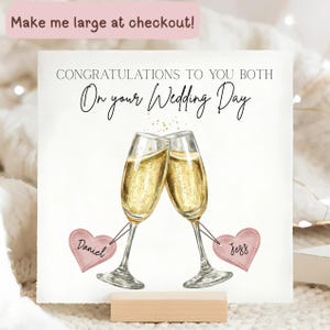 Pode incluir: Cartão de casamento com o texto "CONGRATULATIONS TO YOU BOTH On your Wedding Day". Duas taças de champanhe a brindar, com os nomes "Daniel" e "Jess" em etiquetas de coração rosa. O cartão está num suporte de madeira.