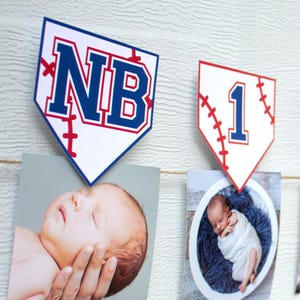 Könnte beinhalten: Eine monatliche Fotogirlande im Baseball-Design für Babys. Die Girlande besteht aus Wimpeln in Form von Baseball-Homeplates mit den Buchstaben "NB" und den Zahlen 1 bis 12. Jede Wimpel enthält ein Babyfoto, das an einer Schnur befestigt ist.